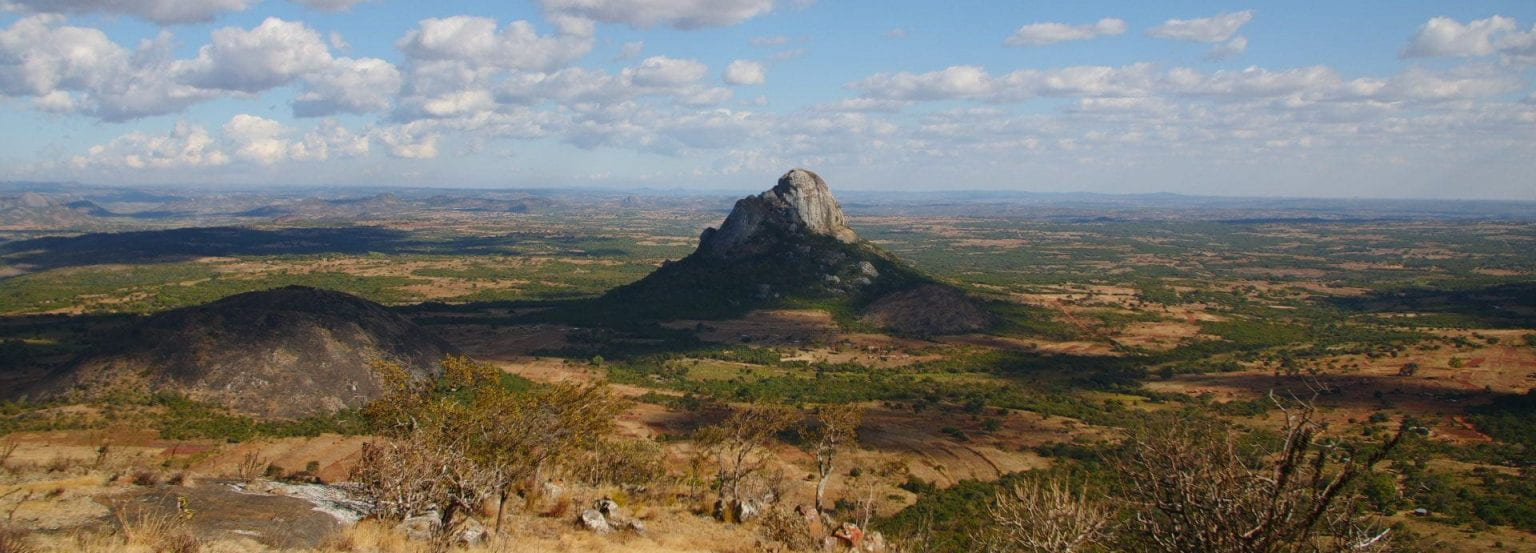 a distant view of a single rocky mountain that juts out abruptly from a plain of grassland and spotted patches of trees. Other than a large nearby hill, most everything else is flat