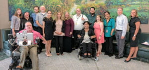 A group of the men and women who work at Tools for Life or for other partner offices. Thirteen people are standing in front of a couch and a large landscape painting, and two are seated in the front in mobility devices. 