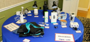 Various assistive technologies are displayed on a round table covered by a blue tablecloth. One sign on the left side of the table reads "Telepresence Devices," and a second sign on the right reads "Wearables." Some of the technologies include fitness and health trackers, specialized clothing, and lamps and door locks controlled by Alexa and Echo devices. 