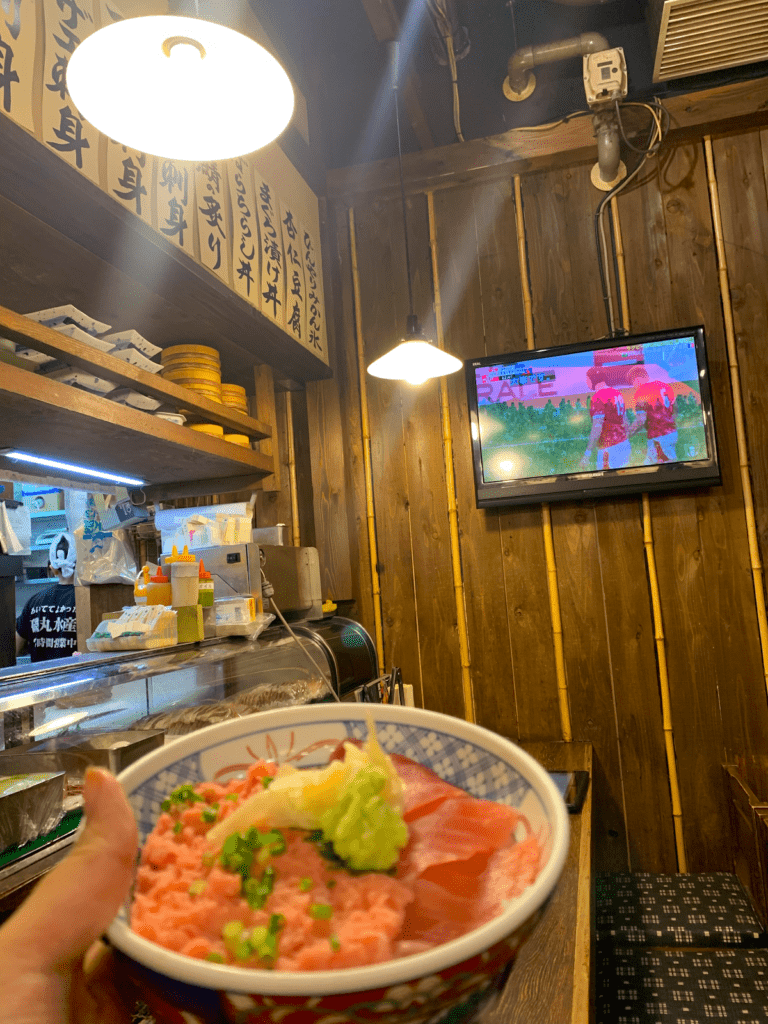 Cy holds up a bowl of fresh tuna in front a TV playing a rugby match