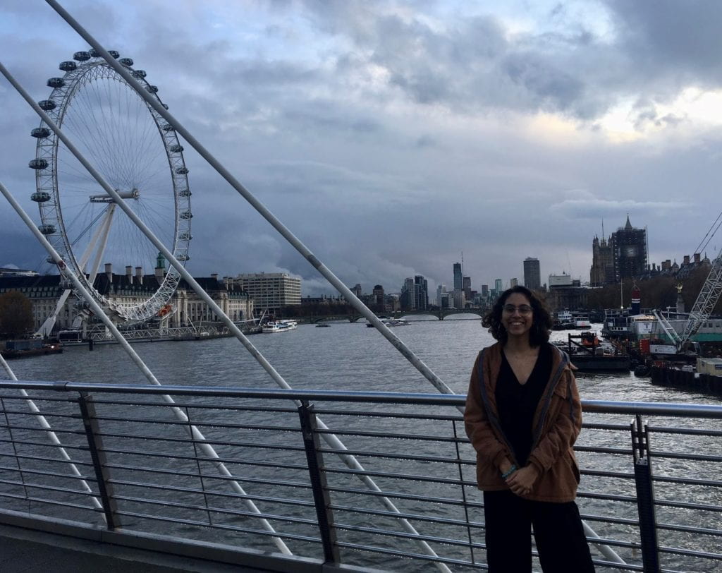 Lea stands on the Hungerford Bridge with the London Eye and Big Ben in the background